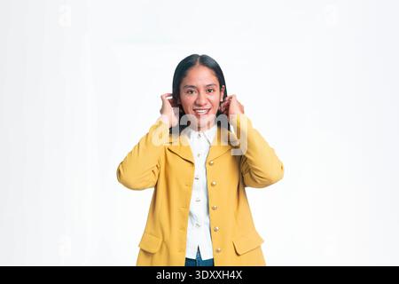 Ritratto di una donna indonesiana felice in un blazer giallo che regola i capelli dietro le orecchie sorridendo alla fotocamera, isolata su sfondo bianco. Foto Stock