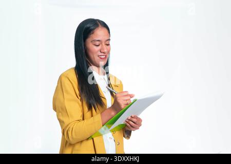 Donna indonesiana professionista in blazer giallo che guarda in basso e scrive su un portadocumenti verde con una penna, isolata su sfondo bianco studio. Foto Stock
