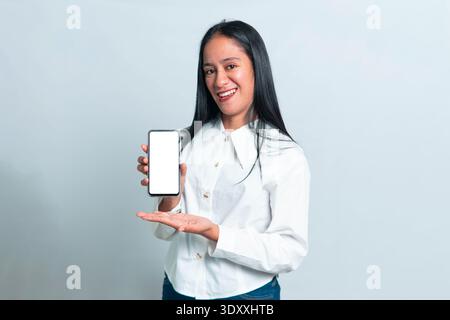 Ritratto di una donna indonesiana sorridente con una camicia bianca professionale pulita che mostra un telefono cellulare con uno schermo bianco luminoso, isolato sul bianco Foto Stock