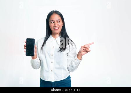 Ritratto di una donna indonesiana sorridente con una camicia bianca professionale che tiene un telefono cellulare con uno schermo nero vuoto e punta il dito verso la S. Foto Stock