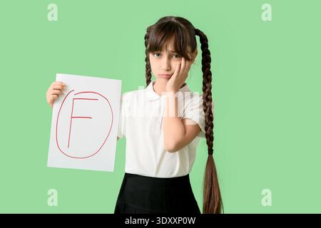 Triste studentessa che tiene carta con risultati negativi del test scolastico su sfondo verde Foto Stock