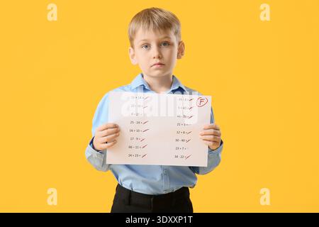 Scolaro triste che tiene carta con risultati negativi del test scolastico su sfondo giallo Foto Stock