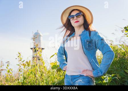 Donna elegante con giacca in denim, occhiali da sole e cappello da sole, posa sicura all'aperto. Ripresa ad angolo basso in un campo verde con un faro sfocato su un sole Foto Stock