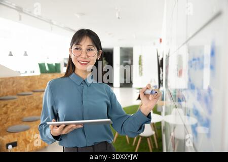 Giovane donna d'affari asiatica positiva in occhiali che lavora sul piano di progetto Foto Stock