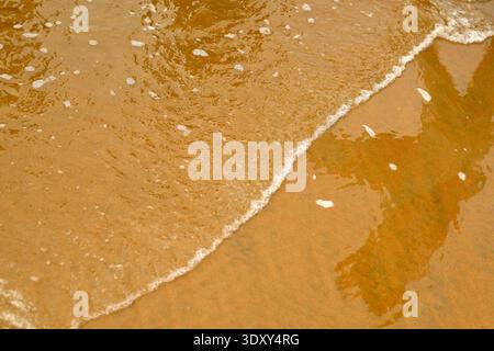 Le onde bagnano delicatamente la spiaggia di sabbia. Una dolce onda oceanica con schiuma bianca su una spiaggia sabbiosa. Foto Stock