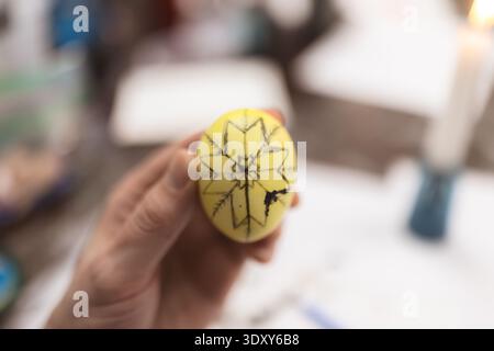 Le mani tengono in mano un uovo giallo con un intricato design a stella nera durante una lezione della comunità per la creazione di un pisanky ucraino. Foto Stock