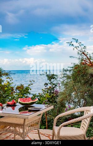 Terrazza estiva con mobili in vimini e anguria a fette sul tavolo, affacciata su un bellissimo lago di montagna. Cespugli spinosi. Foto Stock