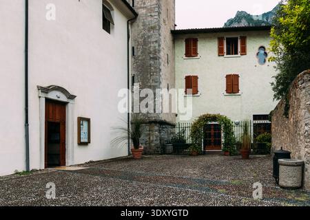 Un incantevole cortile della chiesa a Limone sul Garda presenta edifici in pietra, persiane in legno e un terreno acciottolato con vista sulle montagne. Foto Stock