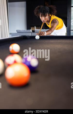 Donna afroamericana che si schiera sul tavolo da biliardo in feltro nero nella sala giochi con il top giallo Foto Stock