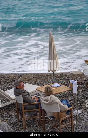 Coppia che gusta vino rosato su una spiaggia di ciottoli a Nizza, Francia. Onde mediterranee, ombrellone chiuso, ambiente di viaggio rilassato. Foto Stock