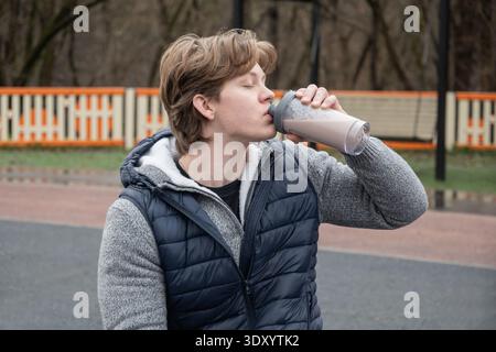Un giovane uomo siede su una panchina da palestra all'aperto che regge un frullato proteico e indossa un gilet casual. Si rilassò, godendosi un momento di riposo durante l'allenamento. Foto Stock
