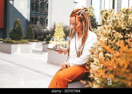 Una donna moderna di mezza età con dreadlock guarda lo schermo dello smartphone. La donna usa il telefono per strada. Donna moderna in città || modello rilasciato Foto Stock
