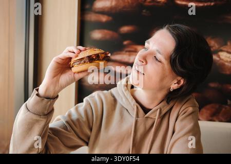 Giovane donna che tiene in mano un hamburger di manzo con pancetta su un panino di sesamo mentre siede in un ristorante fast food e guarda fuori dalla finestra. Concetto di casual Foto Stock