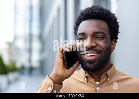 Giovane adulto sorridente molto, tenendo un telefono cellulare all'orecchio, ascoltando una conversazione e comunicando mentre si trova in un ambiente urbano moderno Foto Stock