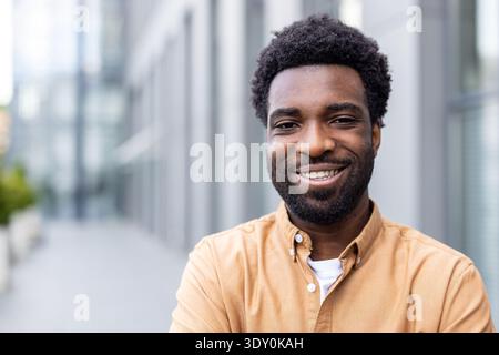 Giovane e fiducioso africano che sorride brillantemente mentre si trova all'aperto in un ambiente cittadino, esprimendo felicità e successo con un atteggiamento amichevole e accogliente Foto Stock