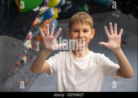 Uno scolaro felice con una t-shirt bianca si trova in una palestra di arrampicata mostrando le sue palme aperte ricoperte di polvere di magnesio bianco. Concetto di sport infantile suc Foto Stock