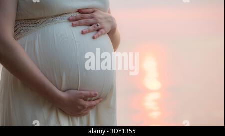Una donna incinta è in piedi vicino al mare sul retro del suo letto, da vicino, accarezzandosi la pancia Foto Stock