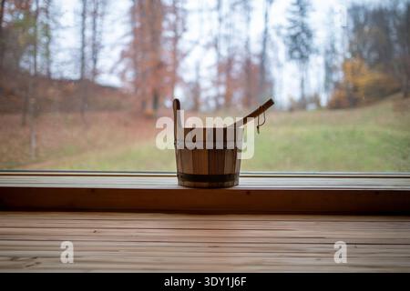 Un secchio di legno e un mestolo d'acqua poggiano su una panchina di legno liscia all'interno di una sauna. La benna è cilindrica con spranghe verticali in legno e una fascia metallica. A l Foto Stock
