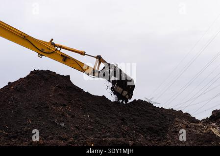 La benna dell'escavatore scarica il terreno scuro, mostrando le operazioni di movimento terra e i lavori di costruzione Foto Stock