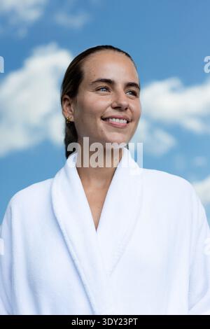 Donna ventenne che posa all'aperto sotto il cielo blu, indossa un accappatoio bianco e cerchi dorati. Ritratto, luce del giorno, aria aperta, nuvole, sereno, minimalista, Foto Stock