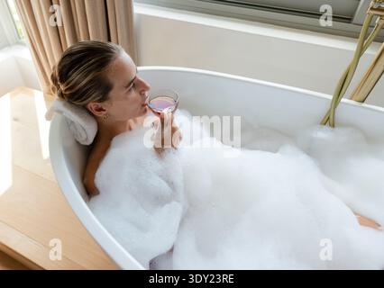 Donna adulta sdraiata nella vasca da bagno indipendente con bolle, tenendo il vetro con stelo, appoggiando la testa su un asciugamano. Donna, bagno, schiuma, rosa, lusso, serena, finestra Foto Stock