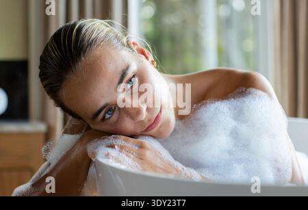 Donna della metà degli anni '20 sdraiata nella vasca da bagno con schiuma bianca, che riposa il mento sul bordo, che guarda la finestra. Vasca da bagno, indipendente, minimalista, serena, luce naturale, Foto Stock