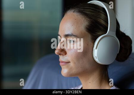 Donna che indossa cuffie bianche sopra l'orecchio accanto alla finestra con un cuscinetto blu-grigio rivolto verso destra. Ritratto, calma, contemplativa, ascolto, musica, natu Foto Stock