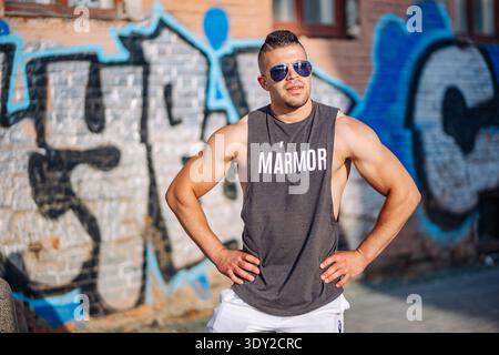 Mosca, Russia - 25 agosto 2025: Culturista muscoloso che indossa occhiali da sole, canotta, posa con sicurezza accanto al muro dei graffiti in un ambiente urbano russo Foto Stock