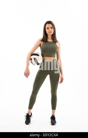 Una giovane donna tiene la pallavolo, sta in piedi con fiducia. L'abbigliamento sportivo verde si adatta perfettamente e mostra movimento. Le sneaker nere contrastano con la tavolozza dei colori tenui. SH Foto Stock