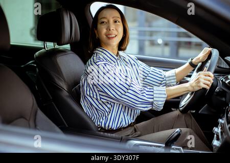 Una donna coreana siede al posto di guida di un'auto. Sorride e tiene il volante. L'ambiente appare moderno e al chiuso. Sembra che sia giorno Foto Stock