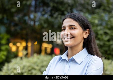 Giovane donna d'affari indiana che sorride e guarda lontano, si gode una tranquilla pausa all'aperto in un parco cittadino, sicura di sé e rinfrescata durante una pausa di lavoro diurna Foto Stock