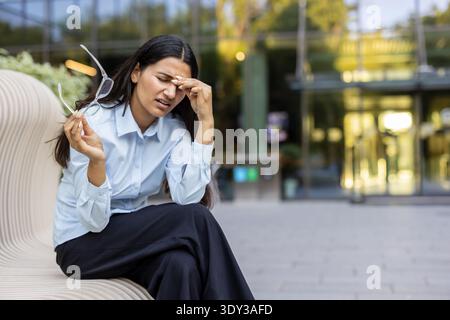 Giovane donna d'affari con occhiali in mano che soffre di mal di testa doloroso e affaticamento degli occhi, seduta su una panchina mentre si sente stressata e bruciata dal lavoro Foto Stock