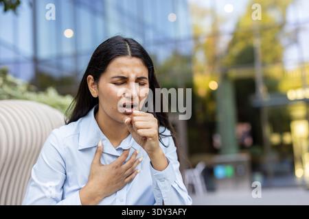 Giovane donna affetta da tosse dolorosa e fastidio al petto, tenendo la mano alla bocca e al petto mentre si sente male in un ambiente moderno all'aperto Foto Stock