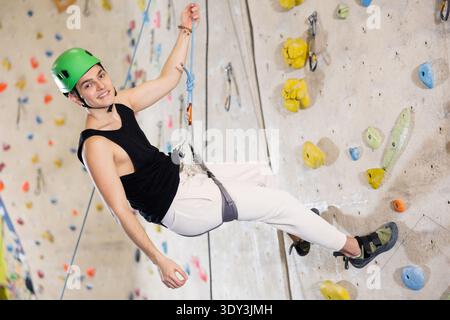l'uomo con casco di sicurezza e con la fune di sicurezza si appende vicino alla parete di arrampicata in palestra Foto Stock
