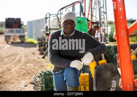 Sorridente contadino afroamericano in piedi all'aperto appoggiandosi sulla macchina agricola Foto Stock