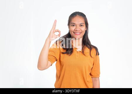 Felice donna indonesiana con camicia arancione che mostra un buon segno con la mano, sorridendo alla telecamera su uno sfondo bianco pulito. Foto Stock