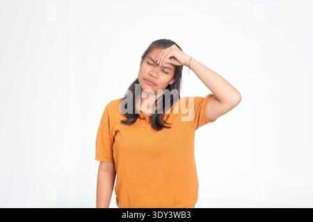 Giovane donna indonesiana in camicia arancione che sembra debole e malata, toccando la fronte mentre soffre di vertigini o mal di testa. Foto Stock