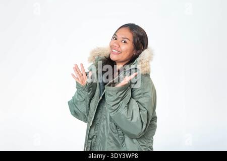 Una allegra donna del sud-est asiatico con un caldo cappotto invernale verde oliva con cappuccio in pelliccia sorridente su sfondo bianco. Foto Stock