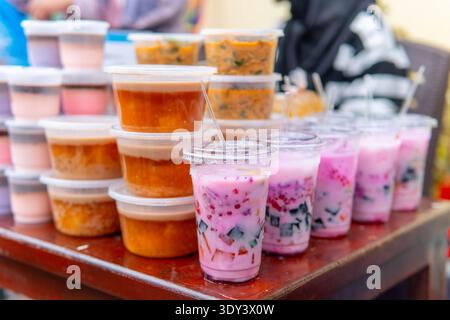 Vari spuntini tradizionali indonesiani in contenitori di plastica e cocktail di frutta ghiacciata colorata su un tavolo di legno in un mercato di Street food per iftar Foto Stock