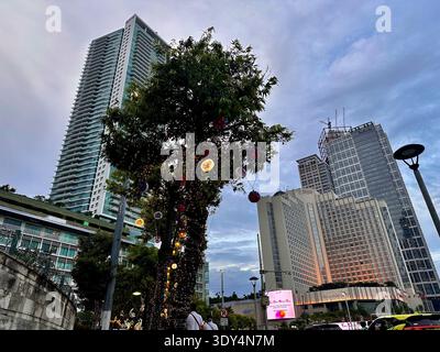 Giacarta - 5 marzo 2026: Un albero avvolto da luci sorge accanto a una strada trafficata e grattacieli al tramonto nel centro di Giacarta. Foto Stock