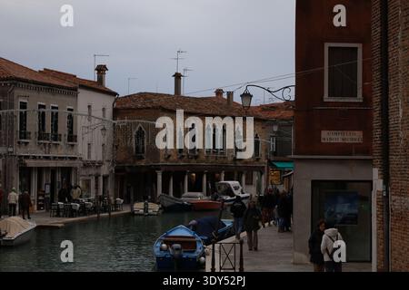 Canale fiancheggiato da piccole barche ed edifici storici, dove i pedoni camminano lungo la stretta strada sul lungomare sotto un cielo coperto. Foto Stock