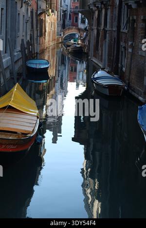 Vista ravvicinata di un tranquillo canale veneziano con piccole barche riflesse nell'acqua immobile sotto edifici storici. Foto Stock
