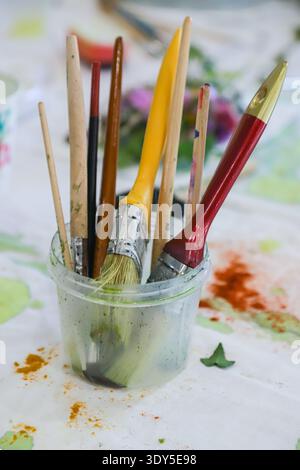 Vari pennelli in un contenitore si trovano su un tavolo adornato da spruzzi di vernice colorata e pezzi di verde che catturano l'essenza di un artista Foto Stock
