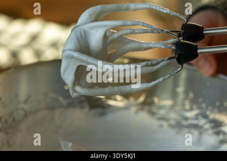 Primo piano di meringa lucida spessa che reggono picchi rigidi sulle frullatrici sopra un recipiente in acciaio inossidabile. Foto Stock