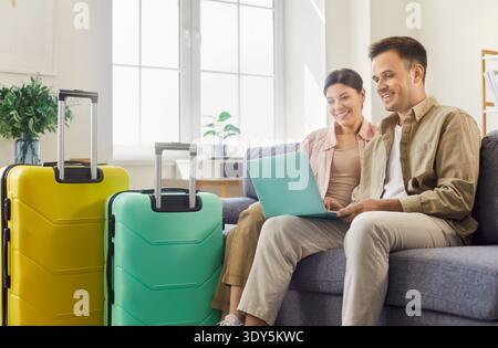 Coppia felice che pianifica una vacanza online a casa con valigie, prenotando tramite laptop Foto Stock