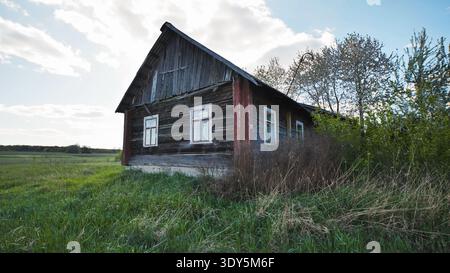 Vecchia casa in legno abbandonata nel villaggio slavo circondata da erba verde e alberi Foto Stock