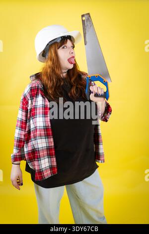 Donna che indossa un elmetto che tiene la sega che fa la faccia divertente Foto Stock