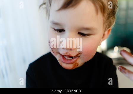 Primo piano ritratto di un bambino di tre anni con cioccolato spalmato su tutta la faccia. Disordine appiccicoso dopo aver infranto le regole della dieta. Concetto di alimentazione malsana, allergie, diabete e obesità infantile. Foto Stock