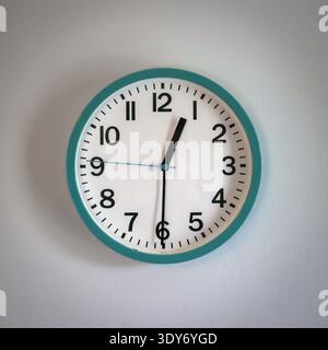 Un orologio da parete turchese a buon mercato con quadrante bianco che mostra le lancette alle dodici e trenta, appeso al muro in una casa. Foto Stock