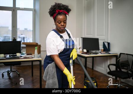 Giovane donna adulta nera che pulisce il pavimento dell'ufficio con un aspirapolvere che indossa guanti e tuta da lavoro, in piedi vicino a scrivanie e monitor di computer in un moderno spazio di lavoro Foto Stock